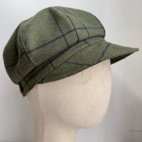 A green tweed flat cap with a blue plaid pattern, displayed on a beige fabric mannequin head against a plain white background.