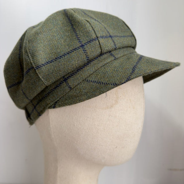 A green tweed flat cap with a blue plaid pattern, displayed on a beige fabric mannequin head against a plain white background.