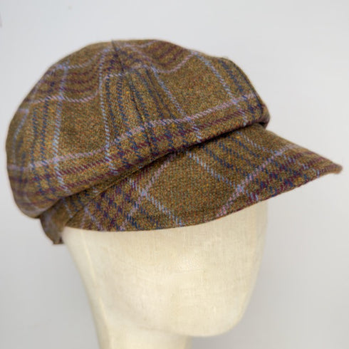 A brown tweed flat cap with a plaid pattern in purple and beige, displayed on a cream-colored mannequin head against a plain white background.