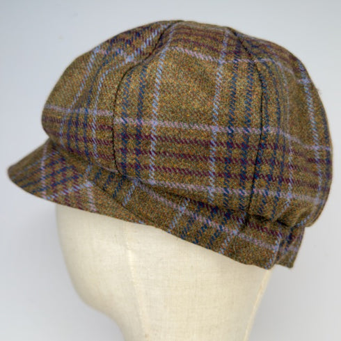 A brown tweed flat cap with a plaid pattern in purple, blue, and beige, displayed on a cream-colored mannequin head against a light gray background.