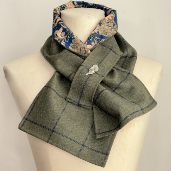 A folded plaid scarf draped on a cream mannequin torso. The main body of the scarf is olive green with a blue windowpane plaid pattern, and the inner lining features a floral print in blue, beige, and cream.