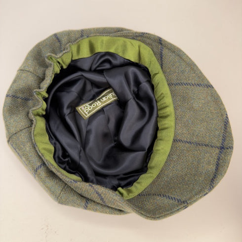 A top-down view of a classic flat cap in a green and blue plaid tweed pattern, featuring a dark navy satin interior and a green fabric trim around the crown.