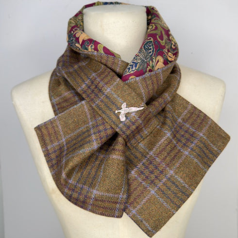 A folded plaid scarf draped on a cream-colored mannequin torso. The scarf features a brown, purple, and beige plaid pattern on the exterior, with a multicolor paisley pattern lining visible at the top edge.