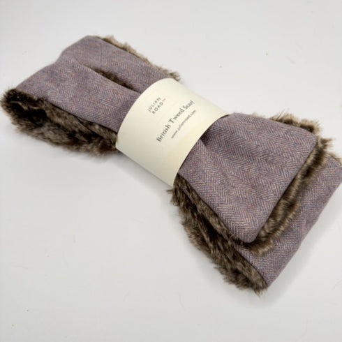 A folded British Tweed scarf in a dusty purple herringbone pattern with a brown faux fur trim, wrapped in a white paper band.